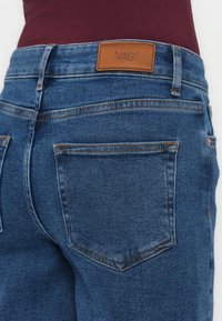Bakfra av person med blå denimjeans med et lærmerket fra YAS på livlinjen og en bordeaux topp som er stappet inn.