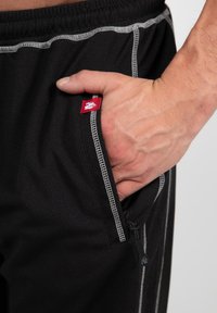 Black athletic pants with a textured fabric, featuring a zippered pocket, contrasting gray stitching, and a red logo tag on the pocket edge.