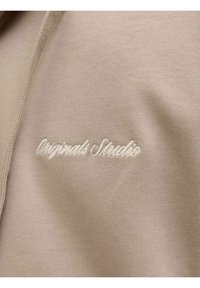 Tissu beige avec texte brodé blanc indiquant « Originals Studio » en écriture cursive sur un matériau doux et tricoté.