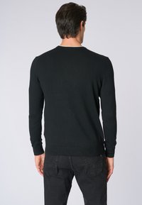Maglione nero a maglia con maniche lunghe, scollatura rotonda e orlo a coste. Il materiale sembra morbido, senza modelli o accentuazioni visibili. Indossato con jeans scuri.