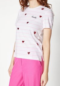 Camiseta de manga corta blanca con rayas rosas, adornada con corazones de lentejuelas rojas y "Voilà" en texto negro; con aberturas en los laterales del dobladillo.