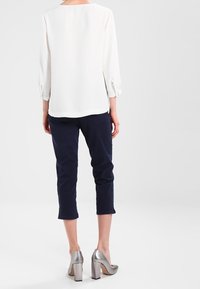 Femme portant une blouse blanche à manches longues, un pantalon cropped bleu marine et des chaussures à talons carrés argentées, se tenant face à un fond gris clair.