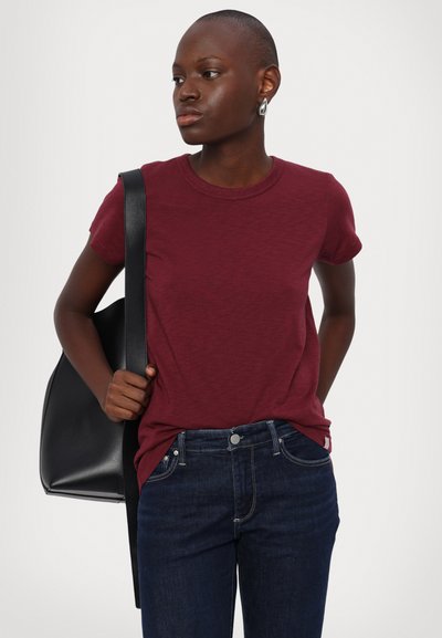 Burgundy crew-neck t-shirt with short sleeves, made of soft fabric, paired with dark blue jeans; model holds a black leather bag.