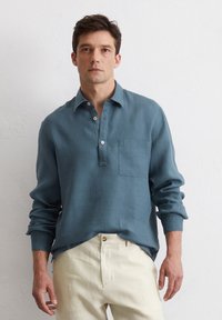 Blue long-sleeve shirt made of lightweight fabric, featuring a collar, a half-button placket, and a left chest pocket. Paired with light-colored pants.