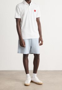 Polo en coton blanc avec un logo patch rouge, associé à un short bleu clair à fines rayures, des baskets blanches et des chaussettes blanches.