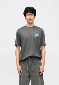 T-shirt de algodão cinzenta com mangas curtas e decote redondo. Apresenta texto gráfico em verde-petróleo no lado esquerdo do peito. Combinada com calças cinzentas com cordão.