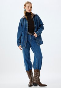 Denimjacka med brunt läderkrage, tillsammans med blå denimbyxor och bruna cowboyboots med intrikata sömmar och spetsiga tår.