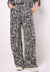 Pantaloni a gamba larga con strisce zebrate bianche e nere, indossati con le mani in tasca e sneakers beige su uno sfondo semplice.