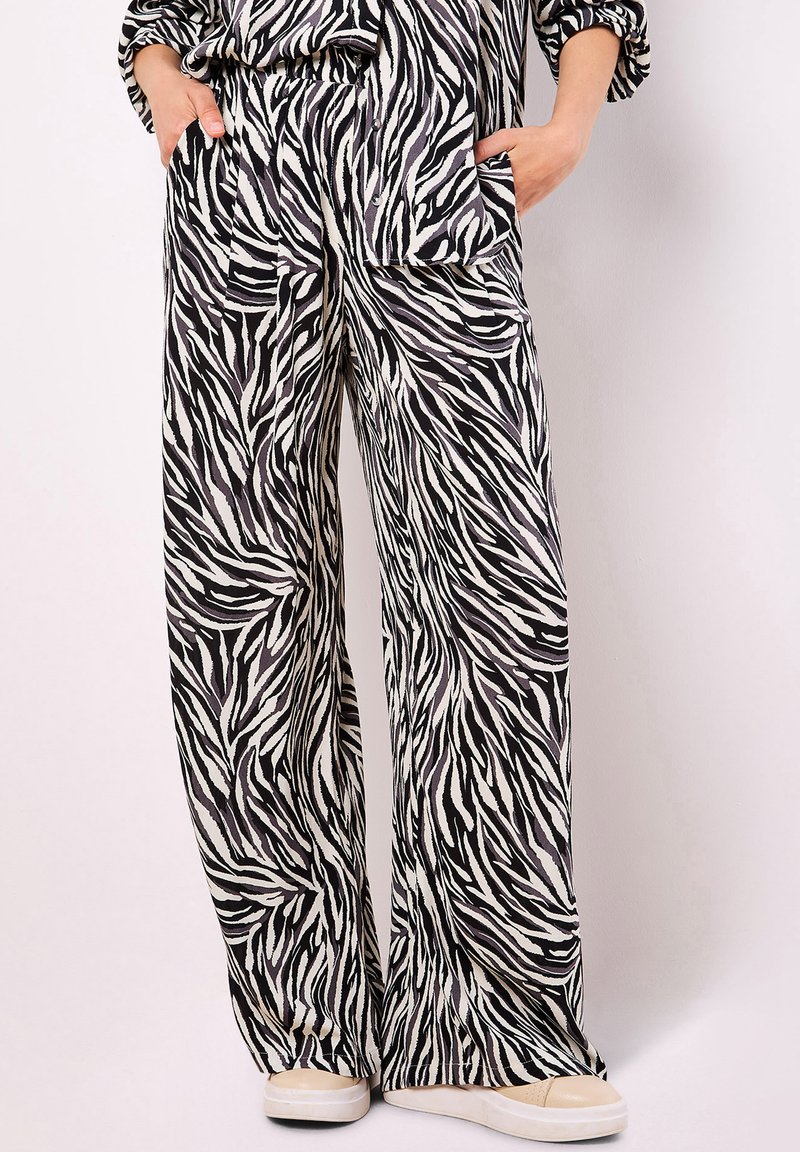 Pantaloni a gamba larga con strisce zebrate bianche e nere, indossati con le mani in tasca e sneakers beige su uno sfondo semplice.