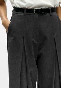 Sive krojene hlače iz teksturirane tkanine, z ostrimi sprednjimi gubami in stranskimi žepi, v kombinaciji z črnim usnjenim pasom.
