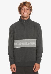 Pullover in pile grigio con colletto alto, zip a quarto e una fascia grigia a contrasto con il logo "Quiksilver" in bianco. Texture morbida e vestibilità comoda.