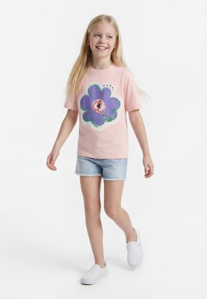 Młoda dziewczyna idzie, ubrana w różowy t-shirt z dużym fioletowym kwiatowym wzorem, jasne jeansowe szorty i białe buty wsuwane.