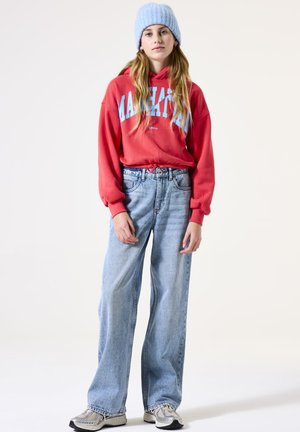Jeune femme se tenant debout, portant un hoodie rouge, un bonnet bleu clair, un jean taille haute à jambes larges et des baskets grises, devant un fond clair.