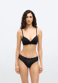 Γυναίκα με μακριά κατσαρά μαλλιά που φοράει μαύρο σουτιέν με λεπτές τιράντες και μαύρο δαντελωτό εσώρουχο, στέκεται μπροστά σε απλό φωτεινό φόντο.