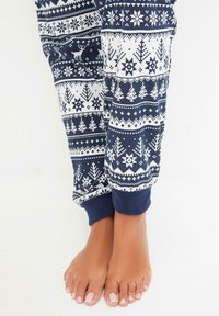 Pantalons de pyjama navy avec des motifs de flocons de neige et d'arbres blancs. Caractéristiques des poignets élastiques. Pieds nus avec un vernis à ongles neutre visibles à l'ourlet.