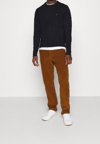 Pull en maille bleu marine, t-shirt gris clair à col rond en dessous, pantalon en velours côtelé marron et baskets blanches. Le mannequin se tient devant un fond uni.