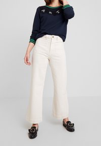 Námořnicky modrý top s dlouhým rukávem, límec s zelenými pruhy a ozdobou "ART"; krémov�é široké kalhoty; černé boty s kruhovým kováním.