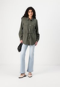 Olijfgroene blouse met knopen en twee borstzakken, getailleerde taille, gedragen met lichtblauwe flared jeans en beige schoenen, terwijl ze een zwarte tas vasthoudt.