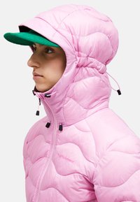 Giacca puffa rosa chiaro con colletto alto e cappuccio attaccato. Presenta una chiusura a zip e dettagli in metallo nero. Cappello verde indossato sotto.
