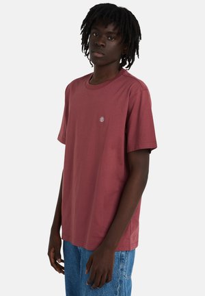 Bordowa bawełniana koszulka z okrągłym dekoltem i krótkimi rękawami, z małym okrągłym logo na piersi. Do tego jasnoniebieskie dżinsy.