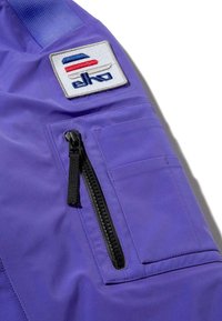 Lila dzsekiszakasz cipzáras zsebbel, piros, fehér és kék részletekkel hímzett logófolttal és texturált szövettel.