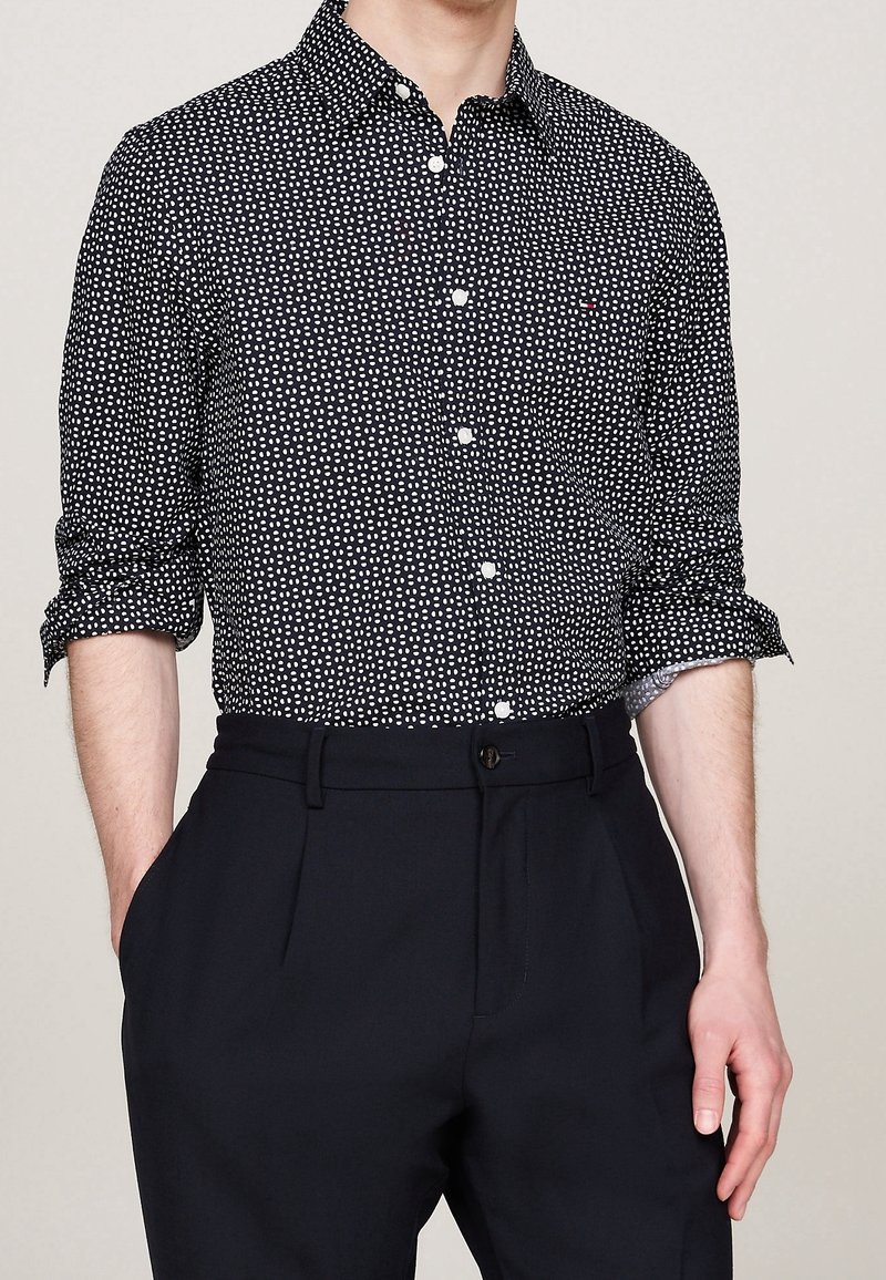 Homme portant une chemise noire à manches longues avec un motif à pois blancs, manches retroussées, rentrée dans un pantalon noir taille haute avec la main dans la poche.