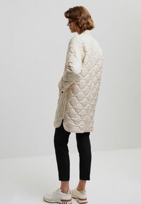 Jasnobeżowy pikowany płaszcz o długim fasonie, z gładką teksturą i bocznymi kieszeniami, zestawiony z czarnymi spodniami i białymi sneakersami.