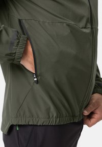 Veste légère verte avec une poche zippée, des poignets élastiques et un ourlet cintré pour un ajustement confortable. Texture de tissu lisse et mate.