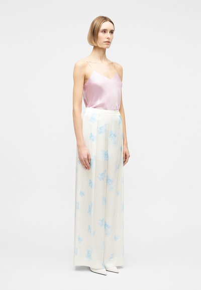 Γυναίκα που στέκεται φορώντας ένα ροζ σατέν τοπ, λευκό παντελόνι με φαρδιά μανίκια και ψηλή μέση με μπλε λουλουδάτα σχέδια, και λευκά μυτερά παπούτσια.