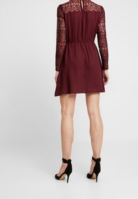 Robe bordeaux avec manches longues en dentelle et taille cintrée, dotée d'une ouverture en forme de clé dans le dos. Portée avec des talons à bride cheville noirs. Texture lisse.