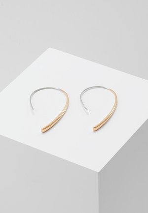 Paire de boucles d'oreilles créoles ouvertes minimalistes bicolores en or et argent reposant sur une surface cubique blanche.
