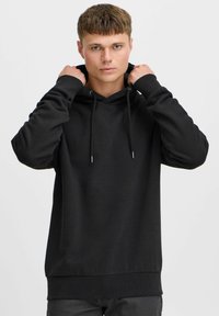 Sudadera con capucha negra hecha de material suave. Presenta cordones, puños acanalados y un dobladillo recto. El modelo se coloca la capucha.