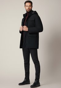 Czarna kurtka puchowa z kapturem, z wieloma kieszeniami z przodu i eleganckim wykończeniem. Nosi ją osoba w bordowym golfie i czarnych spodniach, do których założyła botki za kostkę.