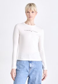 Bež pamukasta majica dugih rukava s okruglim izrezom i crnim logotipom "TOMMYJEANS" na prsima, u kombinaciji s svijetloplavim trapericama.