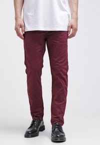 Pantalon bordeaux fuselé en tissu léger froissé avec poches latérales, porté avec un t-shirt blanc et des chaussures noires à lacets.