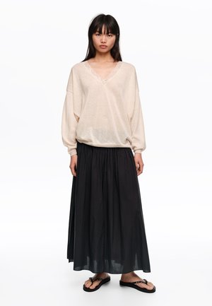 Mujer con un suéter beige holgado de cuello en V con detalle de encaje, una falda larga negra plisada y sandalias negras, de pie contra un fondo blanco.