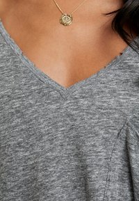 Grauer V-Ausschnitt-Oberteil aus weichem Stoff, mit einer leicht strukturierten Oberfläche, rohen Kanten und einem lockeren Schnitt. Sichtbare goldene Anhänger-Halskette.