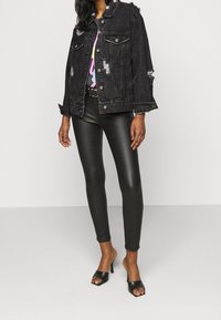 Žena v roztrhané černé džínové bundě, lesklých černých kožených kalhotách, barevném topu a černých volných sandálech na podpatku stojí na jednobarevném pozadí.