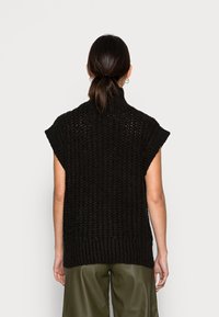 Czarny dziergany top bez rękawów z wysokim kołnierzem i fakturowanymi wzorami. Tył ma otwarte wzory dzianiny i prążkowany brzeg, zestawiony z zielonymi spodniami.