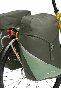 Alforjas impermeables para bicicleta en verde y gris con un diseño triangular, aseguradas por correas. Montadas en una bicicleta con un portaequipajes plateado y un reflector rojo.