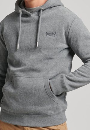 Mann trägt einen grauen Kapuzenpullover mit Kordeln und einer Vordertasche, kombiniert mit hellbraunen Hosen, steht mit einer Hand in der Tasche.