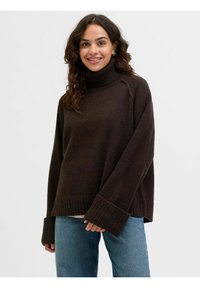 Maglione marrone scuro con collo alto, polsini a coste e vestibilità rilassata. Indossato sopra una maglietta chiara e abbinato a jeans blu.