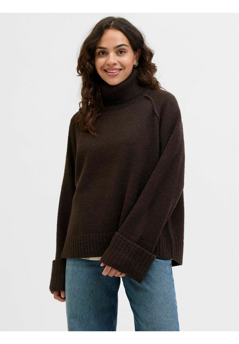 Maglione marrone scuro con collo alto, polsini a coste e vestibilità rilassata. Indossato sopra una maglietta chiara e abbinato a jeans blu.