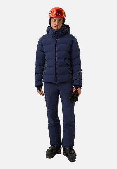 Navy puffer jacket with a hood, zippered pockets, and a fitted design, paired with matching pants and black ski boots. Accessories include gloves.