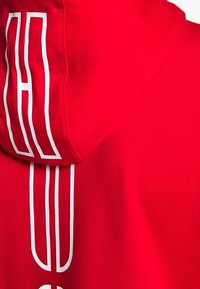 Sudadera roja con una textura suave, que presenta un diseño gráfico blanco distintivo en la parte trasera. La capucha es amplia y no tiene herrajes visibles.