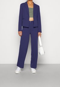 Fialový přizpůsobený blejzr s kapsami na zip, odpovídající široké kalhoty, zelený top a bílá kabelka. Bílé tenisky dokončují celý outfit.