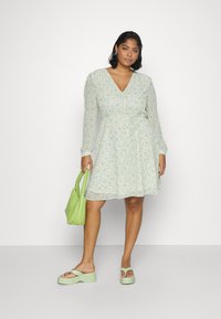 Vestido envolvente floral verde com mangas longas, cintura ajustada e saia rodada. Combinado com sandálias de plataforma verdes e uma bolsa correspondente.