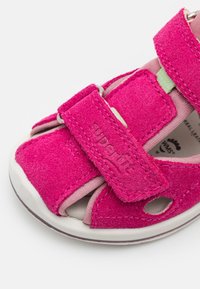 Vaaleanpunaiset mokkasandaalit, joissa on avonainen kärki, säädettävät hihnat ja pehmustettu vuori. Hihnassa on logo ja valkoinen kumipohja.