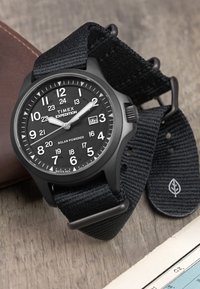 Timex Expedition watch with a black fabric strap, round black case, white markings, solar power feature, and a date window at 3 o'clock.