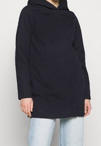 Cappotto con cappuccio di colore blu navy, a taglio dritto, con due tasche frontali e una texture liscia. Chiusura a bottoni coperta e dettagli di cucitura minimi.